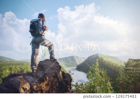 Hiker standing on rocky ledge overlooking river and forest landscape during daytime under blue sky . A female traveler with a backpack on the Mana River and the Manskaya Loop in Siberia, Russia Hiker standing on rocky ledge overlooking river and forest landscape during daytime under blue sky . A female traveler with a backpack on the Mana River and the Manskaya Loop in Siberia, Russia 129175481