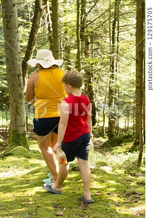 A woman and child hike through a sun-dappled forest, carrying empty water bottles. Enjoying nature's beauty together. 129175705