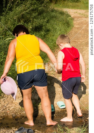 A woman and child walk hand-in-hand along a shallow stream, enjoying a sunny day outdoors. A woman and child walk hand-in-hand along a shallow stream, enjoying a sunny day outdoors. 129175708