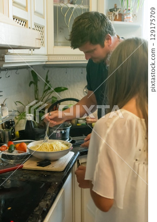 A couple cooks pasta together in their kitchen, sharing a moment of culinary teamwork and domestic bliss. A couple cooks pasta together in their kitchen, sharing a moment of culinary teamwork and domestic bliss. 129175709
