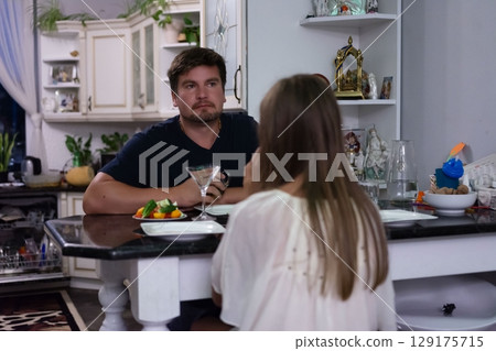 A man and woman sit at a kitchen table, seemingly in an awkward or tense conversation. A small plate of food sits between them. 129175715
