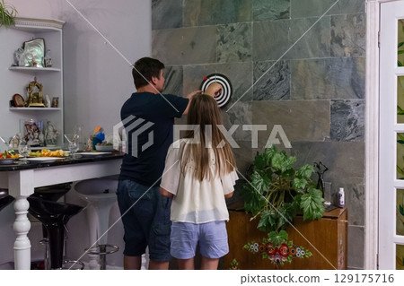 A father and daughter enjoy a game of darts in their home, focusing on their shared activity and the casual atmosphere. A father and daughter enjoy a game of darts in their home, focusing on their shared activity and the casual atmosphere. 129175716