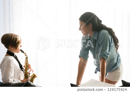 A young student practices the saxophone while receiving guidance from their instructor. A bright, sunlit room provides a calm learning atmosphere. 129175924