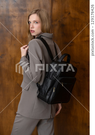 A stylish young woman, long blonde hair, poses in a grey suit, holding a brown leather backpack against a warm wooden background A stylish young woman, long blonde hair, poses in a grey suit, holding a brown leather backpack against a warm wooden background 129176426