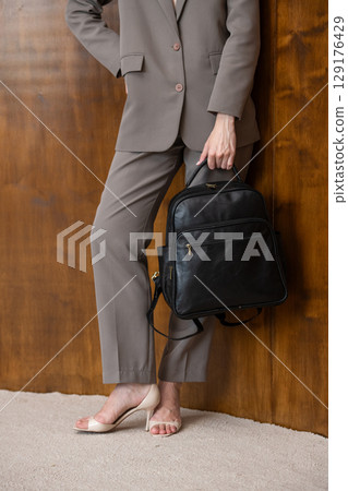 A stylish young woman, poses in a grey suit, holding a brown backpack against a warm wooden background 129176429