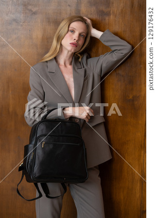 A stylish young woman, long blonde hair, poses in a grey suit, holding a brown leather backpack against a warm wooden background 129176432