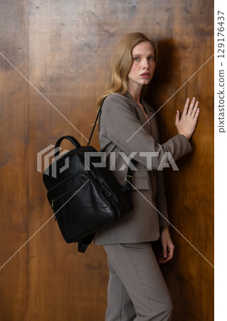 A stylish young woman, long blonde hair, poses in a grey suit, holding a brown leather backpack against a warm wooden background 129176437