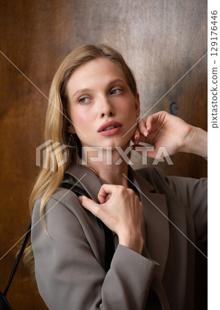 A stylish young woman, long blonde hair, poses in a grey suit, holding a brown leather backpack against a warm wooden background A stylish young woman, long blonde hair, poses in a grey suit, holding a brown leather backpack against a warm wooden background 129176446