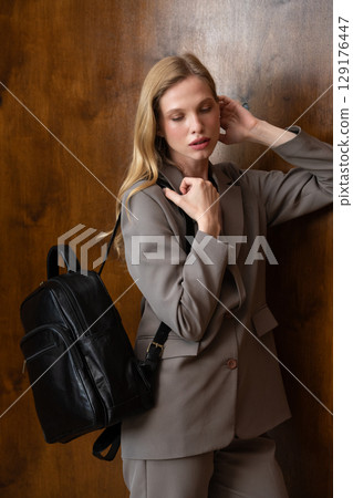 A stylish young woman, long blonde hair, poses in a grey suit, holding a brown leather backpack against a warm wooden background A stylish young woman, long blonde hair, poses in a grey suit, holding a brown leather backpack against a warm wooden background 129176447