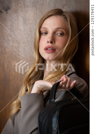 A stylish young woman, long blonde hair, poses in a grey suit, holding a brown leather backpack against a warm wooden background A stylish young woman, long blonde hair, poses in a grey suit, holding a brown leather backpack against a warm wooden background 129176451