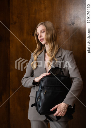 A stylish young woman, long blonde hair, poses in a grey suit, holding a brown leather backpack against a warm wooden background A stylish young woman, long blonde hair, poses in a grey suit, holding a brown leather backpack against a warm wooden background 129176460