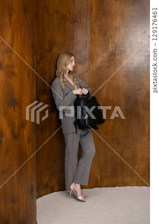 A stylish young woman, long blonde hair, poses in a grey suit, holding a black leather backpack against a warm wooden background A stylish young woman, long blonde hair, poses in a grey suit, holding a black leather backpack against a warm wooden background 129176461