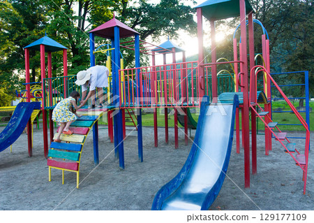 A child receives help climbing a colorful playground structure. Sunlight streams through the trees. 129177109