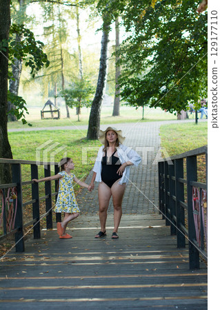 A mother and daughter stroll across a park bridge, the child in a summer dress, the mother in a swimsuit and sun hat. 129177110