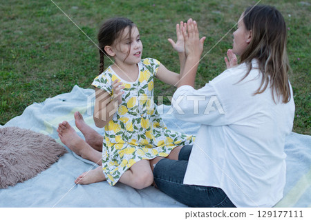 A mother and daughter play a hand-clapping game outdoors on a blanket. 129177111