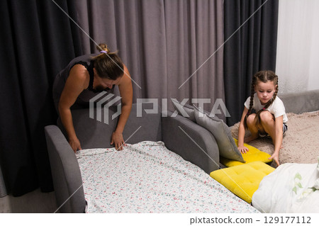 A woman and a girl make a bed together in a room. They are placing a sheet on a sofa bed. 129177112