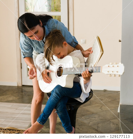 A woman teaches a child to play the guitar, patiently guiding their hands on the strings. Music education and family bonding. 129177113