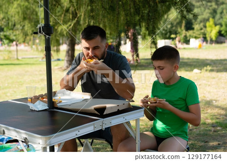 Father and son enjoy a pizza lunch outdoors under a shady tree. 129177164