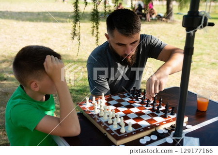 A man and a boy play a game of chess outdoors on a sunny day. Focused concentration on the board. 129177166