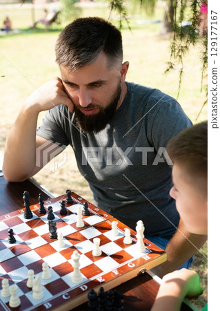 A man and a child play a game of chess outdoors on a sunny day. A man and a child play a game of chess outdoors on a sunny day. 129177167