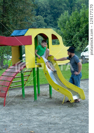 A father helps his son go down a playground slide. They are enjoying a sunny day outdoors. A father helps his son go down a playground slide. They are enjoying a sunny day outdoors. 129177170