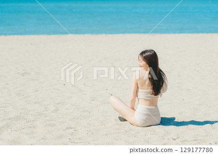 Young woman in swimsuit sitting on sandy beach 129177780