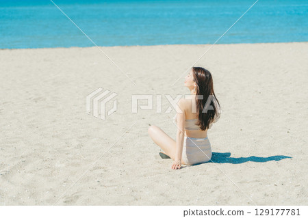 Young woman in swimsuit sitting on sandy beach 129177781