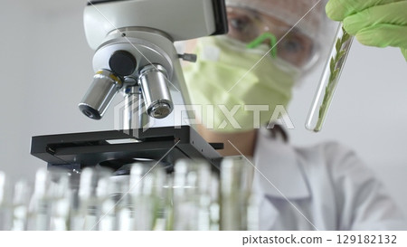 Female scientist wearing green protective mask and gloves is holding test tube with plant sample, researching botany and medical applications using a microscope. Medicine and science concept Female scientist wearing green protective mask and gloves is holding test tube with plant sample, researching botany and medical applications using a microscope. Medicine and science concept 129182132