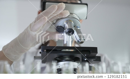 Female scientist wearing white protective gear analyzing test samples under microscope in contemporary research environment, close up of white gloved hand. Medicine and science concept 129182151