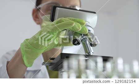 Close up of scientific green gloved hands adjusting microscope lens, blurred laboratory glassware highlighting medical investigation. Medicine, laboratory and science concept Close up of scientific green gloved hands adjusting microscope lens, blurred laboratory glassware highlighting medical investigation. Medicine, laboratory and science concept 129182154