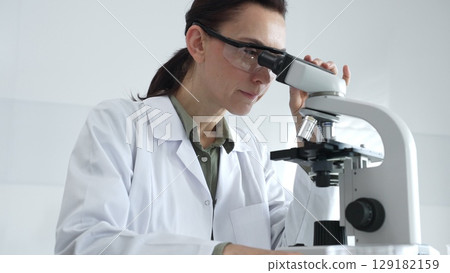 Female scientist in a lab coat and protective glasses examining samples through a microscope in a research laboratory. Medicine, healthcare and science concept 129182159