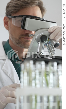Scientific researcher in protective gear analyzing specimens through a microscope in a sterile laboratory environment. Medicine, healthcare and science concept Scientific researcher in protective gear analyzing specimens through a microscope in a sterile laboratory environment. Medicine, healthcare and science concept 129182191