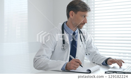 A male doctor in a lab coat with a stethoscope sits at a table in a bright medical office and uses a laptop while taking notes on a clipboard for patient care and treatment A male doctor in a lab coat with a stethoscope sits at a table in a bright medical office and uses a laptop while taking notes on a clipboard for patient care and treatment 129182224