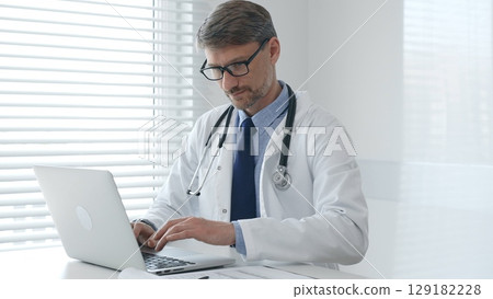 A male doctor in glasses and a lab coat with a stethoscope is typing on a laptop keyboard while sitting at a table in a bright medical office 129182228