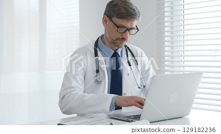 A male doctor in glasses and a lab coat with a stethoscope is typing on a laptop keyboard while sitting at a table in a bright medical office 129182236