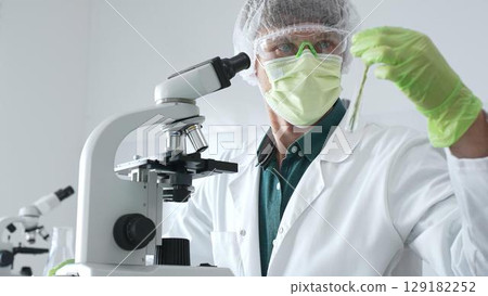 Adult male scientist wearing green protective gear looking at plant samples in test tubes and using a microscope in modern laboratory for botanical research. Medicine, healthcare and science concept 129182252