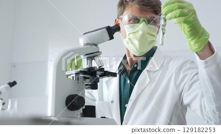 Medical scientist wearing protective mask, cap and green gloves using microscope doing scientific research in laboratory, developing vaccine or medication. Medicine, healthcare and science concept 129182253