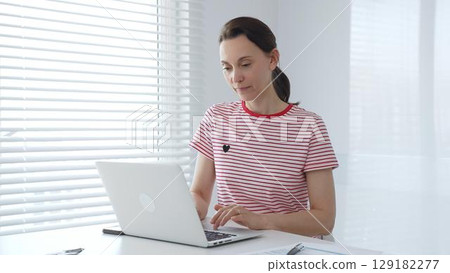 Focused female freelancer working from home, using laptop and sitting at white desk near window with blinds, concentrating on her online work. Business people concept Focused female freelancer working from home, using laptop and sitting at white desk near window with blinds, concentrating on her online work. Business people concept 129182277