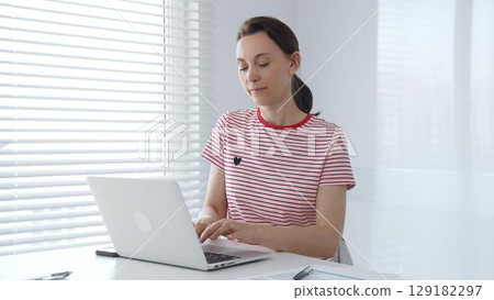 Focused female freelancer working from home, using laptop and sitting at white desk near window with blinds, concentrating on her online work. Business people concept 129182297