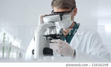 Adult male medical researcher analyzing samples with an advanced microscope, emphasizing safety protocols in a modern laboratory setting. Medicine, healthcare and science concept 129182298