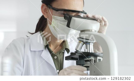 Female scientist examining microscope slide, wearing white protective gear while conducting medical research in laboratory environment. Medicine, health care and science concept 129182302