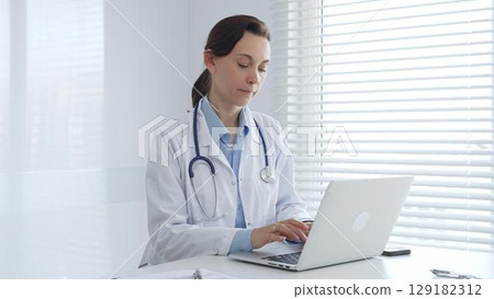 Female doctor working on laptop in white lab coat and stethoscope, sitting at desk in bright, sunlit medical office. Medicine and healthcare concept 129182312