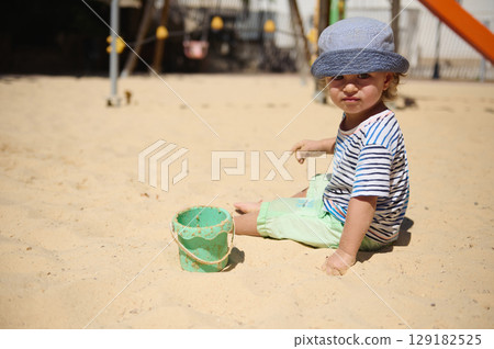 Child Plays in Sandbox with Toy Bucket at Sunny Playground 129182525