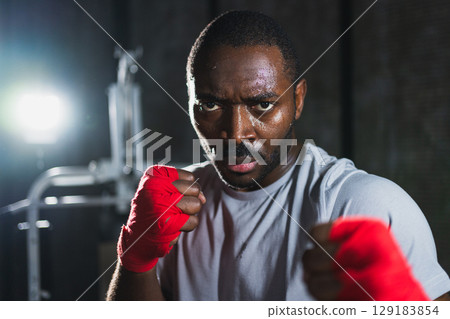 Boxer in gym. Aggressive African man fighter resting after training boxing ready for fight looking at camera. Strong sweated man training punches looking concentrated straight preparing for sparring Boxer in gym. Aggressive African man fighter resting after training boxing ready for fight looking at camera. Strong sweated man training punches looking concentrated straight preparing for sparring 129183854