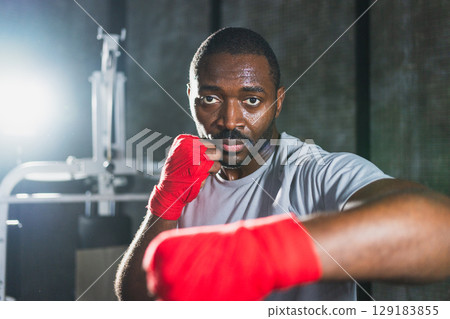 Workout in gym. African man fighter ready for fight punching with boxing wraps protective bandages to camera. Strong man training punches looking concentrated straight preparing for boxing sparring 129183855