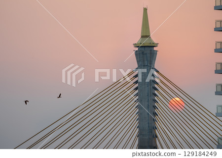 Closeup silhouette of a Bhumibol Bridge with the sun going down over the bridge during orange sunset sky. Closeup silhouette of a Bhumibol Bridge with the sun going down over the bridge during orange sunset sky. 129184249