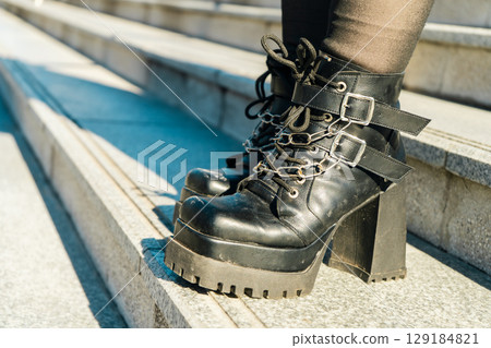 Woman wearing stylish black platform boots with chains and buckles standing on stairs Woman wearing stylish black platform boots with chains and buckles standing on stairs 129184821