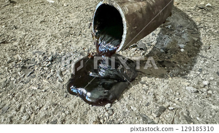 Black oil pouring out of an open pipe on the ground, in a dirty and dusty environment. Black oil pouring out of an open pipe on the ground, in a dirty and dusty environment. 129185310