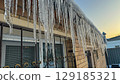 Long glistening icicles hanging from a building roof at sunset, reflecting warm golden light on a cold winter day. 129185321