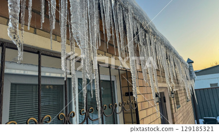 Long glistening icicles hanging from a building roof at sunset, reflecting warm golden light on a cold winter day. Long glistening icicles hanging from a building roof at sunset, reflecting warm golden light on a cold winter day. 129185321